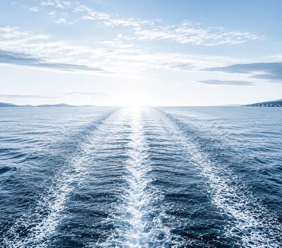 Open ocean view with a boat trail leading toward the horizon, with distant landmasses and a bright sky with scattered clouds.