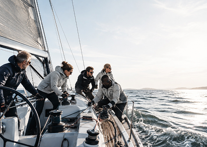 A group of seven people sailing on a boat during daytime, with some crew members adjusting equipment and the ocean in the background.