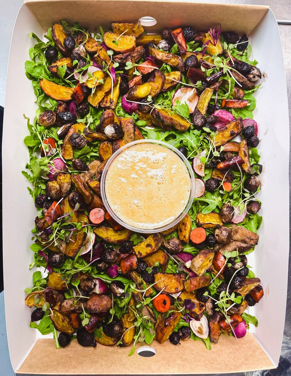 Salad with roasted vegetables like carrots, sweet potatoes, and beets, topped with microgreens, with a small cup of dressing in the center.