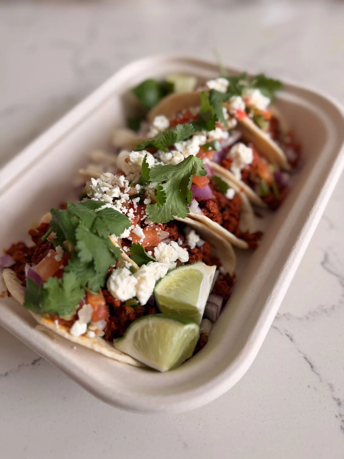 A row of three tacos filled with seasoned meat, topped with cilantro, queso fresco, diced onion, and chopped tomato, garnished with lime wedges, served in a white takeout container.