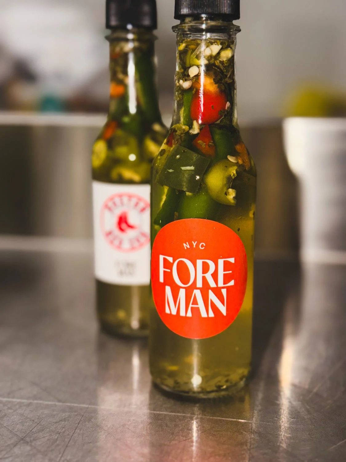 Glass bottle labeled NYC Foreman filled with pickled vegetables including cherry tomatoes, zucchini, and peppers.