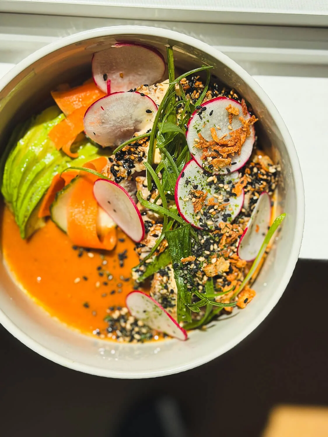 A colorful bowl of poke with sliced radishes, cucumber, carrots, green onions, sesame seeds, and crunchy toppings.