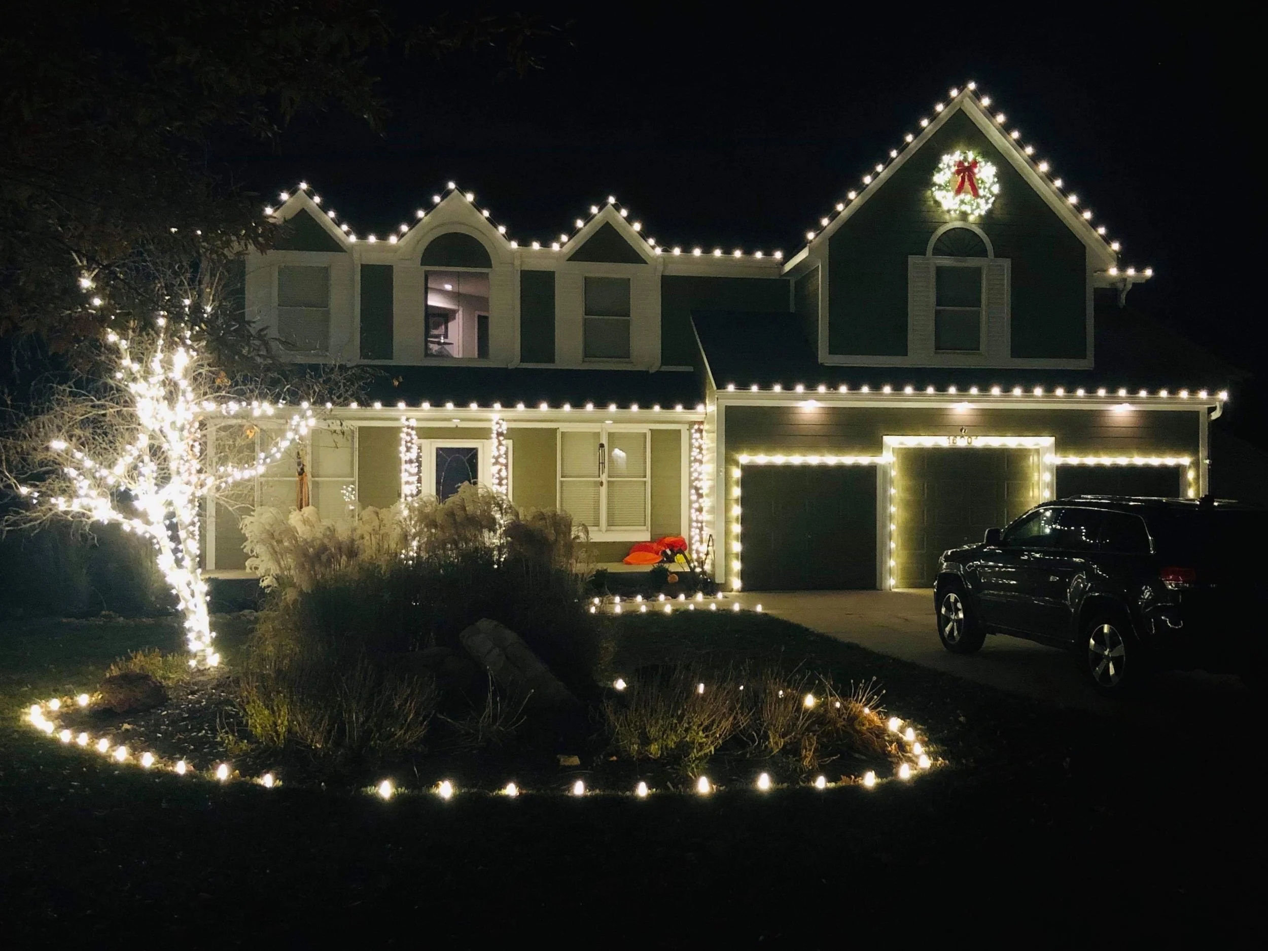 A house decorated with Christmas lights, a wreath with a red bow on the upper right, and a tree wrapped in holiday lights in the front yard at night.