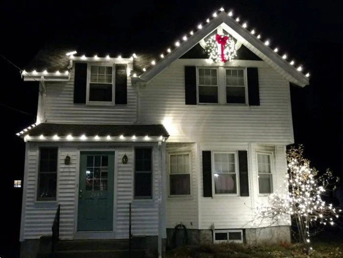 White two-story house decorated with Christmas lights, a wreath on the front facade, and Christmas lights on the trees.