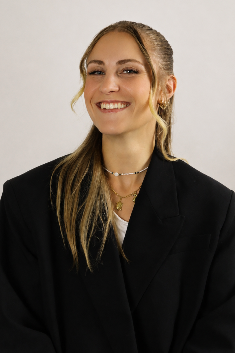 Une jeune femme souriante, cheveux blonde attachés en queue de cheval, portant un blazer noir et des bijoux, pose devant un fond blanc.