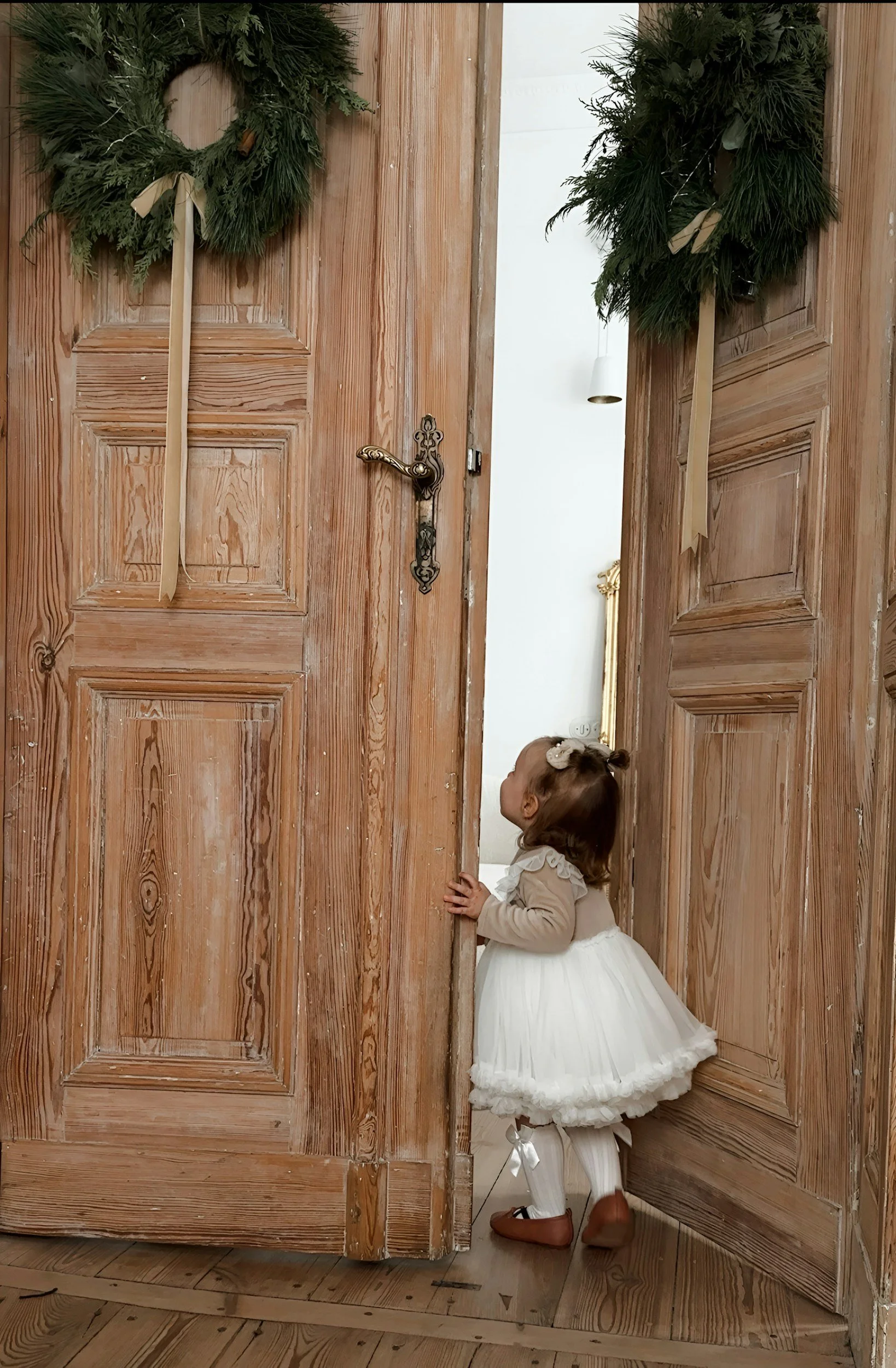 A young girl in a white dress and brown shoes is peeking through an open wooden door decorated with Christmas wreaths. The door has a brass handle and the girl is touching the door with one hand, looking into another room.