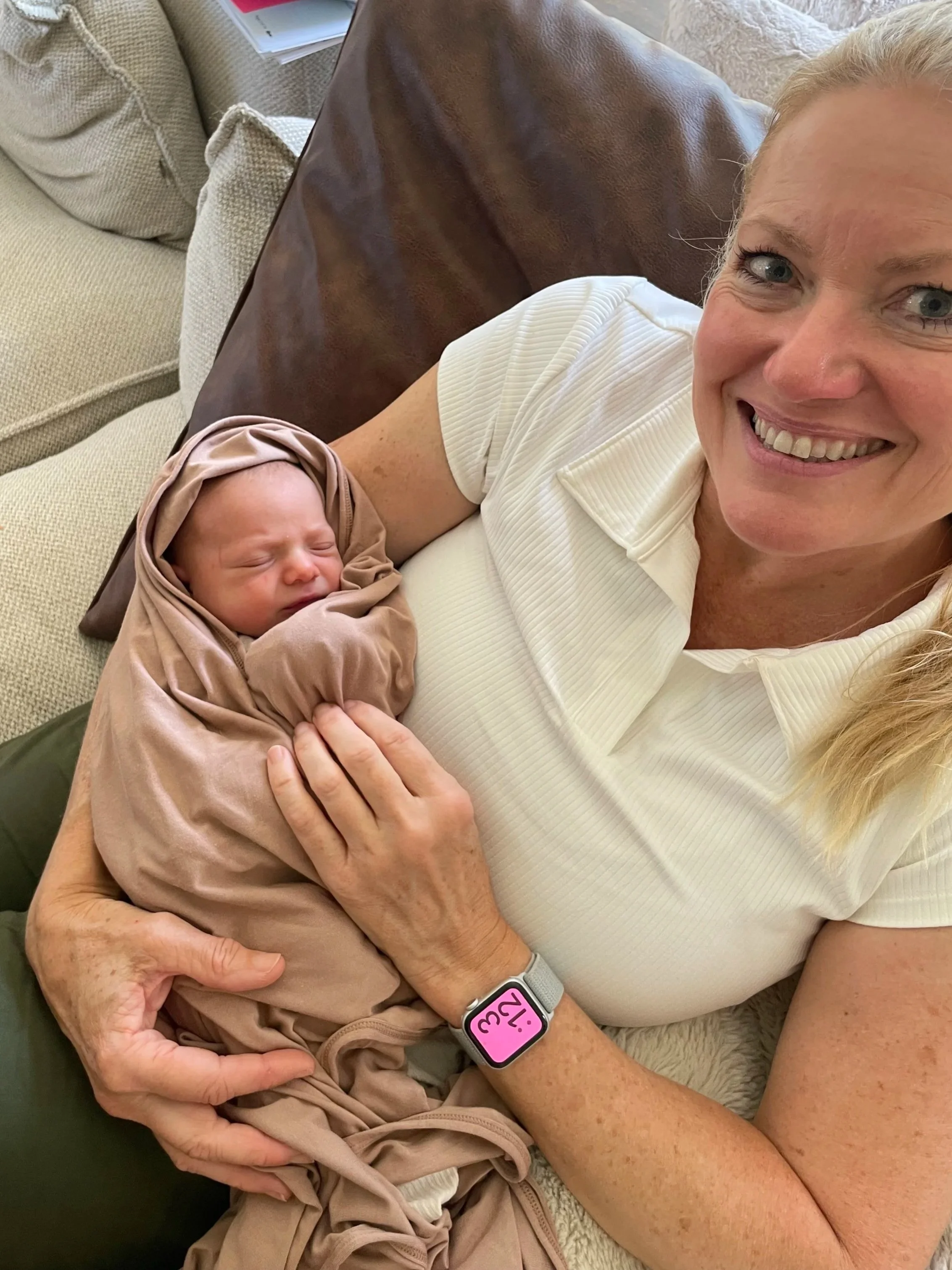 A woman smiling while holding a sleeping newborn wrapped in a beige cloth, sitting on a beige and brown couch.