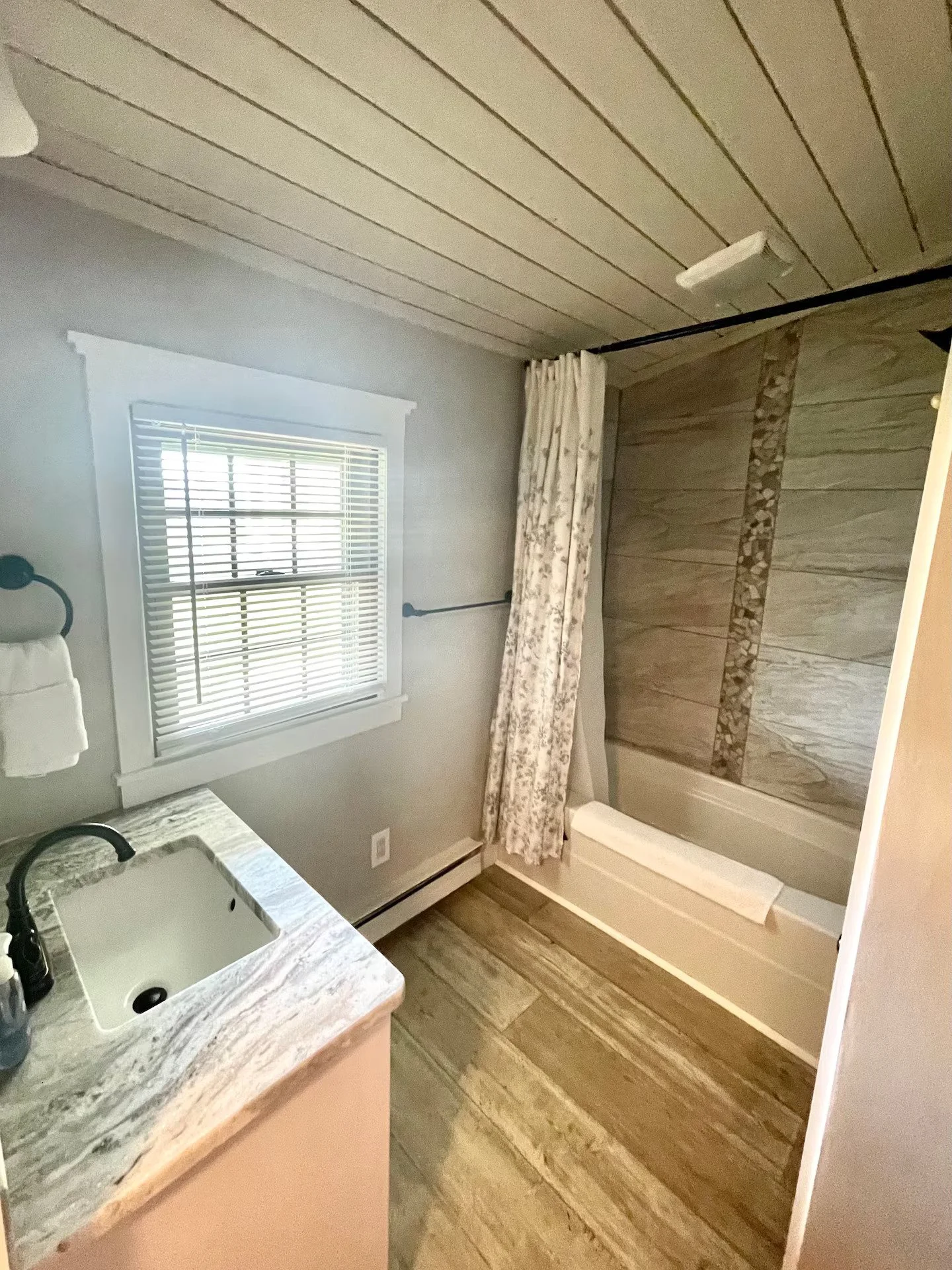 Bathroom with a window, white blinds, a sink with a granite countertop, beige cabinets, a shower with marble tiles and a floral shower curtain, and wood-look flooring.