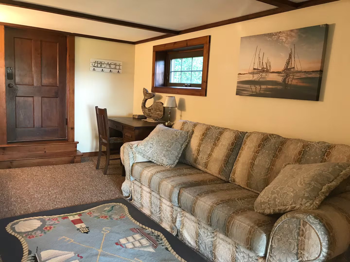 Living room with a patterned beige sofa, a wooden desk with a nautical horse sculpture, and framed artwork of sailboats on the wall. There's a small window and a door with wooden trim, and a colorful decorative rug on the carpeted floor.