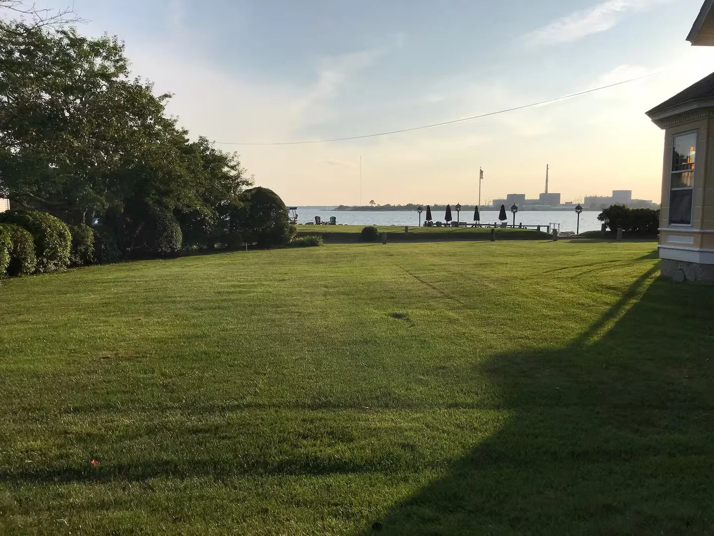 A landscaped backyard with green grass, shrubs, and trees overlooking a body of water. In the distance, there are lounge chairs and umbrellas on a dock, with industrial buildings and an American flag flying on a flagpole. The sky is partly cloudy, and the sun is setting.