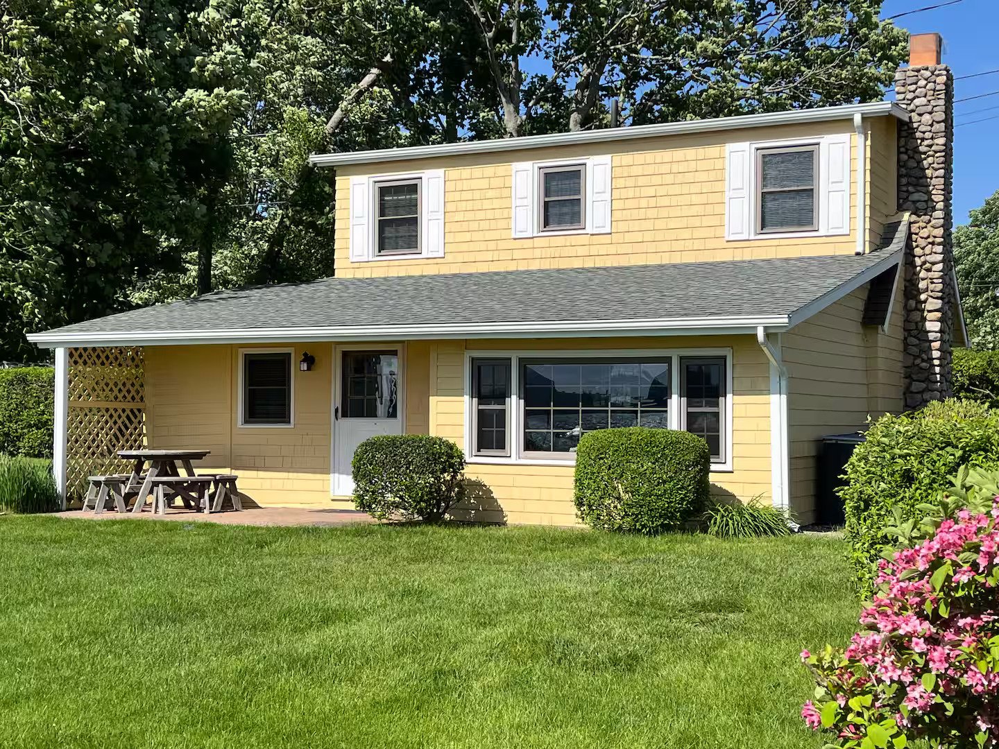Yellow two-story house with white trim, large front window, small front porch with a picnic table, surrounded by green grass, bushes, and trees, with a bright blue sky.