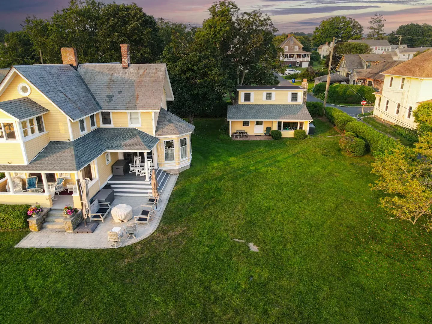 A yellow two-story house with a porch and stairs leading down to a patio, surrounded by a green lawn, and neighboring small yellow and white houses in a residential neighborhood.