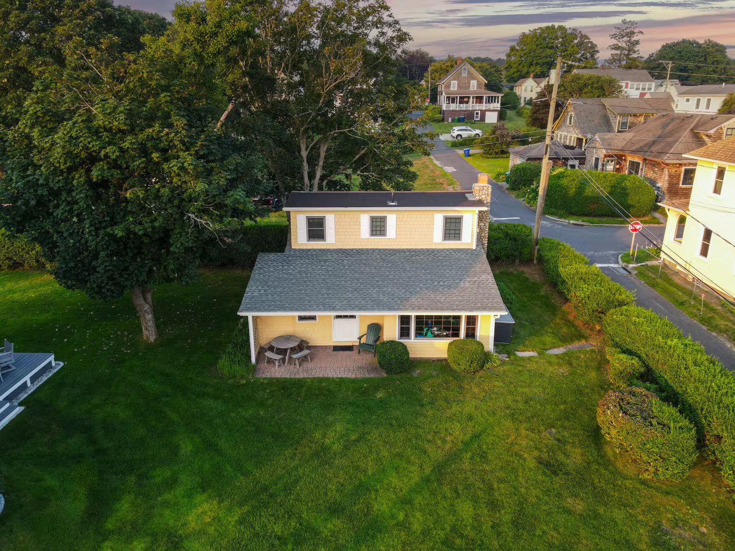 An aerial view of a yellow house with a gray roof, surrounded by a green lawn and bushes, with a large tree on the left, and neighboring houses on the right and background, during sunset.