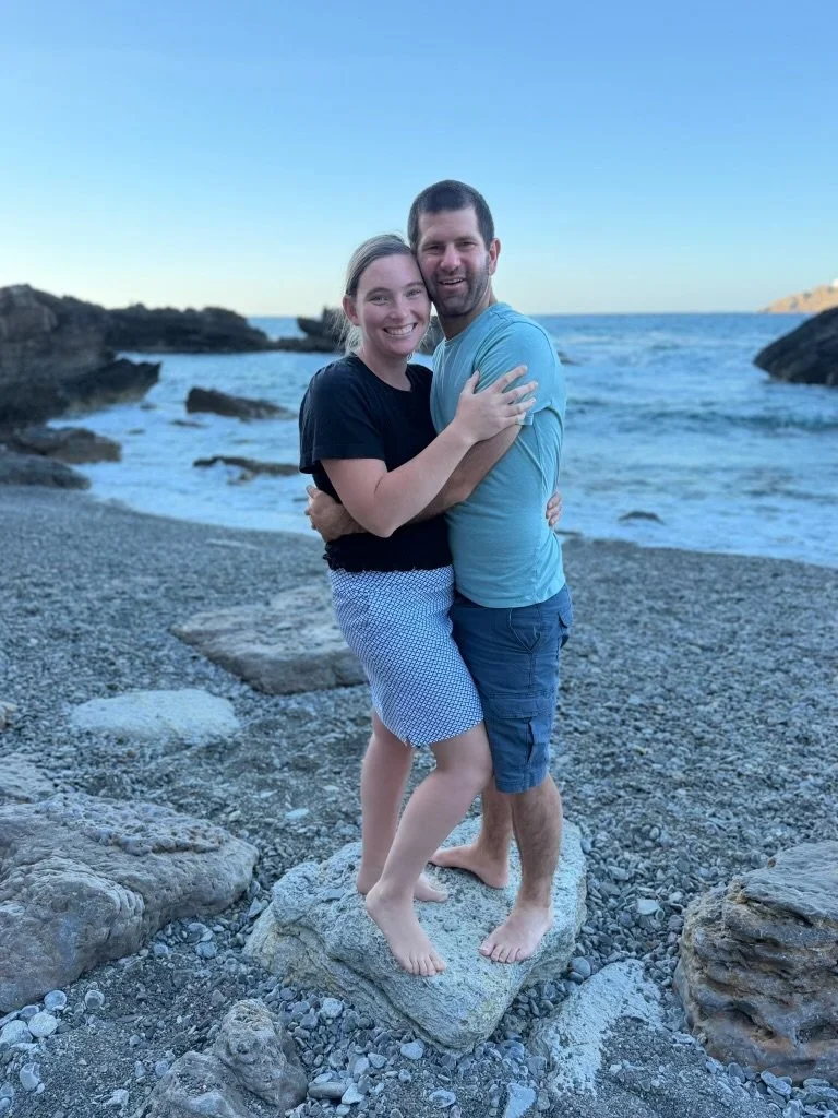 A smiling couple hugging each other on a rocky beach with the ocean and rocks in the background, both standing on a large rock.