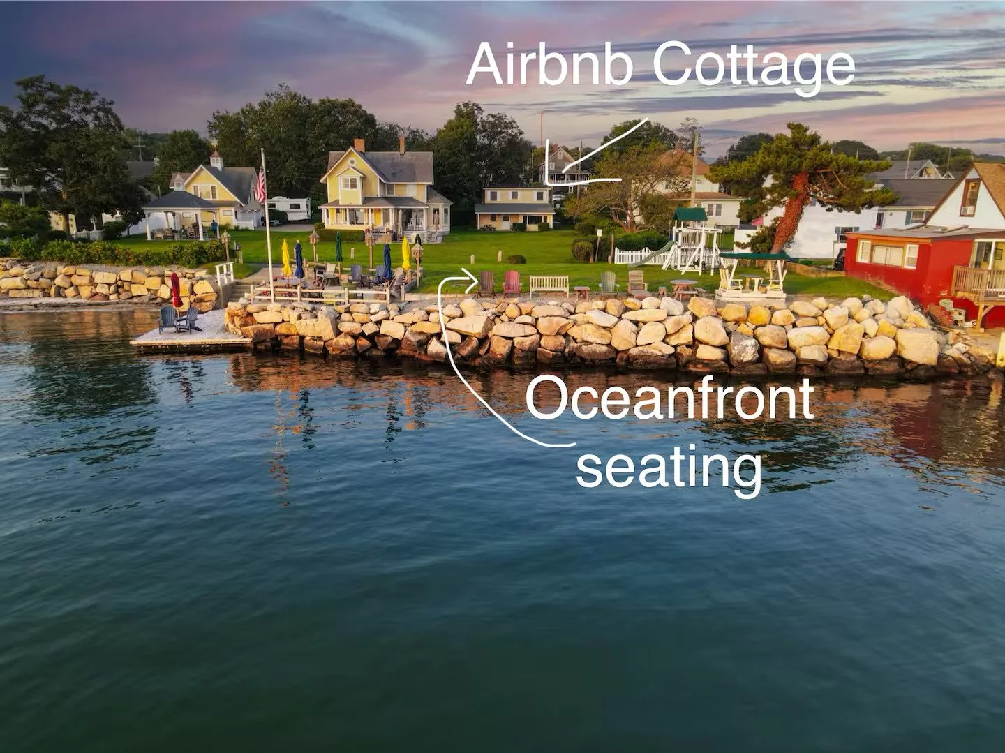 A waterfront scene with a rocky shoreline, oceanfront seating area with chairs and umbrellas, and houses overlooking the water with a sunset sky in the background.