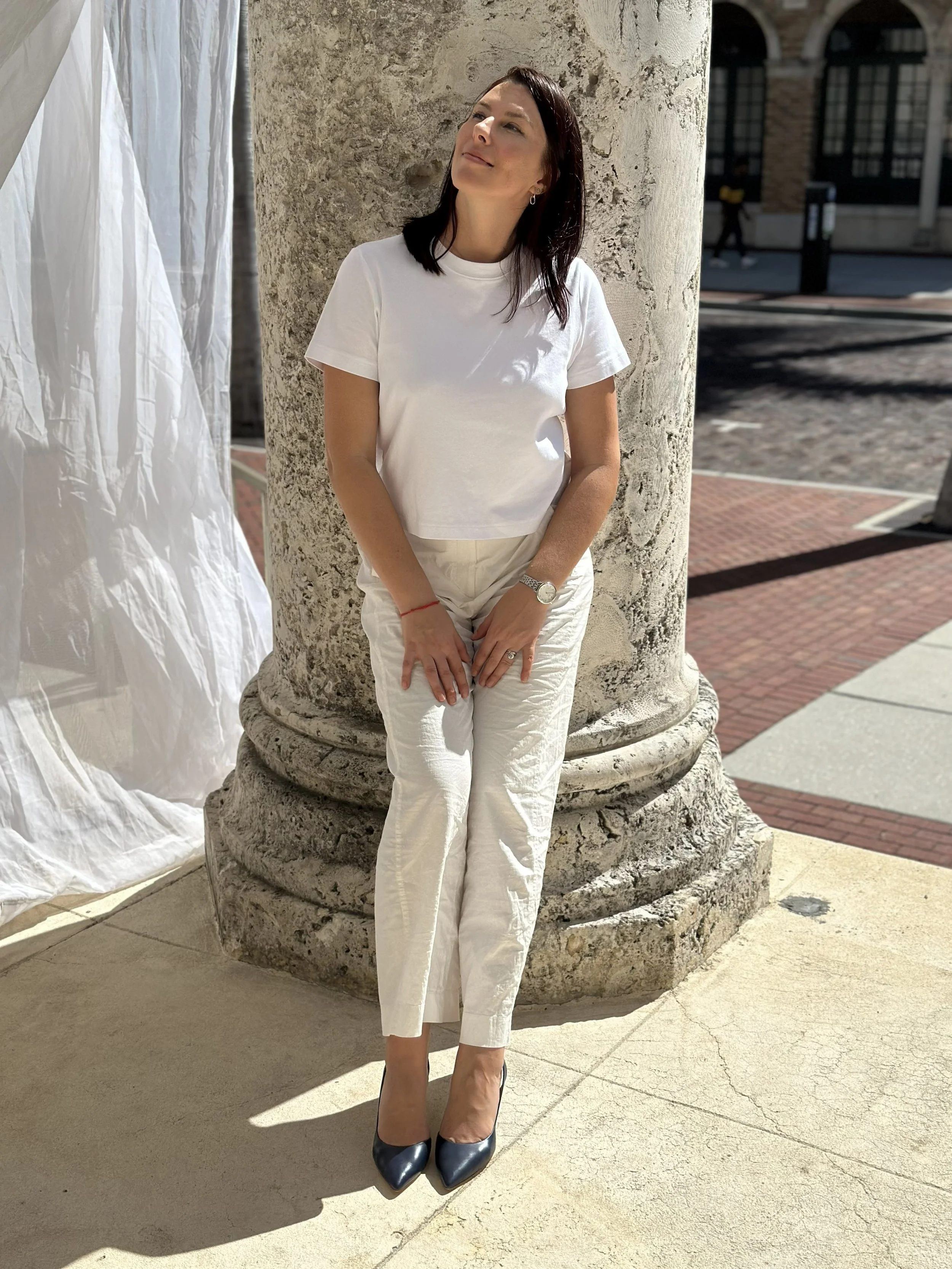 Natallia Khoshchynka Neuro Coach in white t-shirt and white pants standing in the town by the column