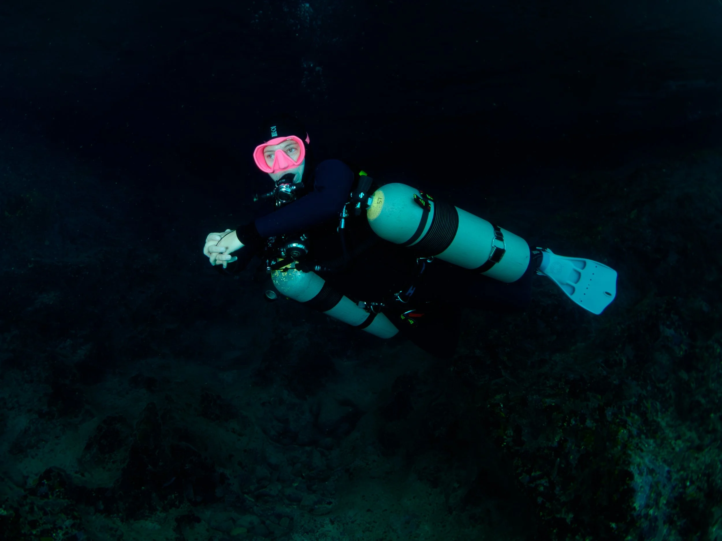 A scuba diver underwater wearing a pink mask and breathing apparatus, surrounded by dark water and rocky ocean floor.