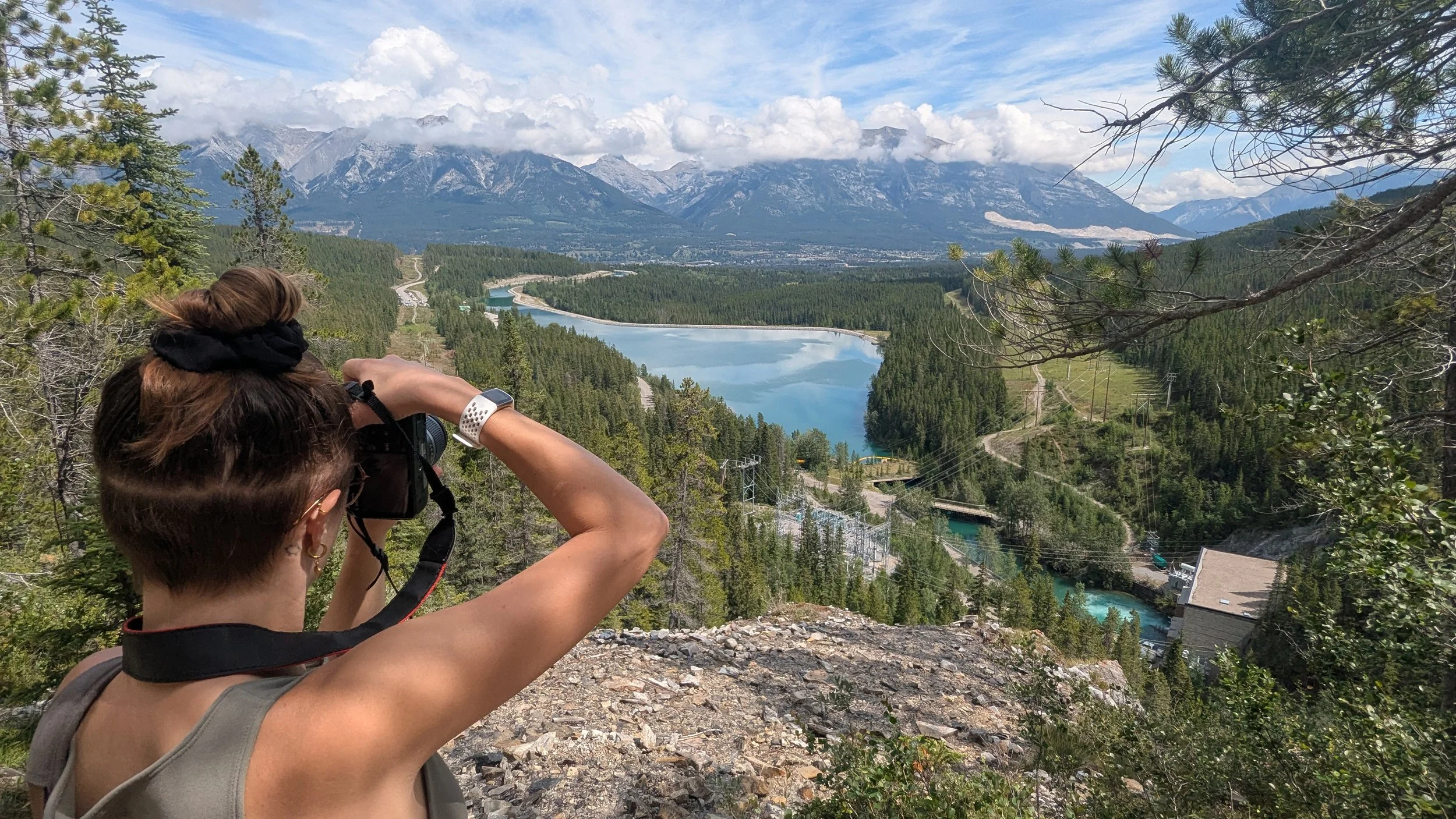 A person taking a photo of a mountainous landscape with a river, trees, and clouds in the background.