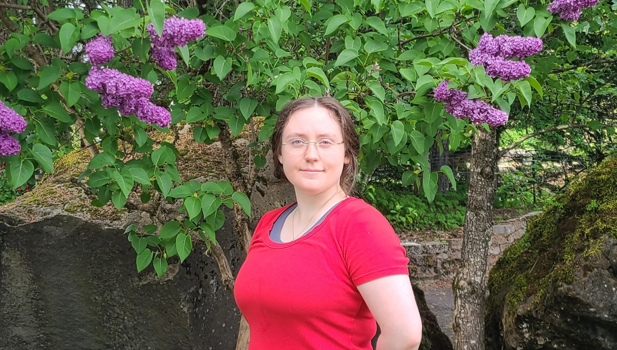 A woman with glasses and wavy brown hair wearing a red shirt and gray undershirt, standing outdoors in front of purple lilac flowers and green foliage.