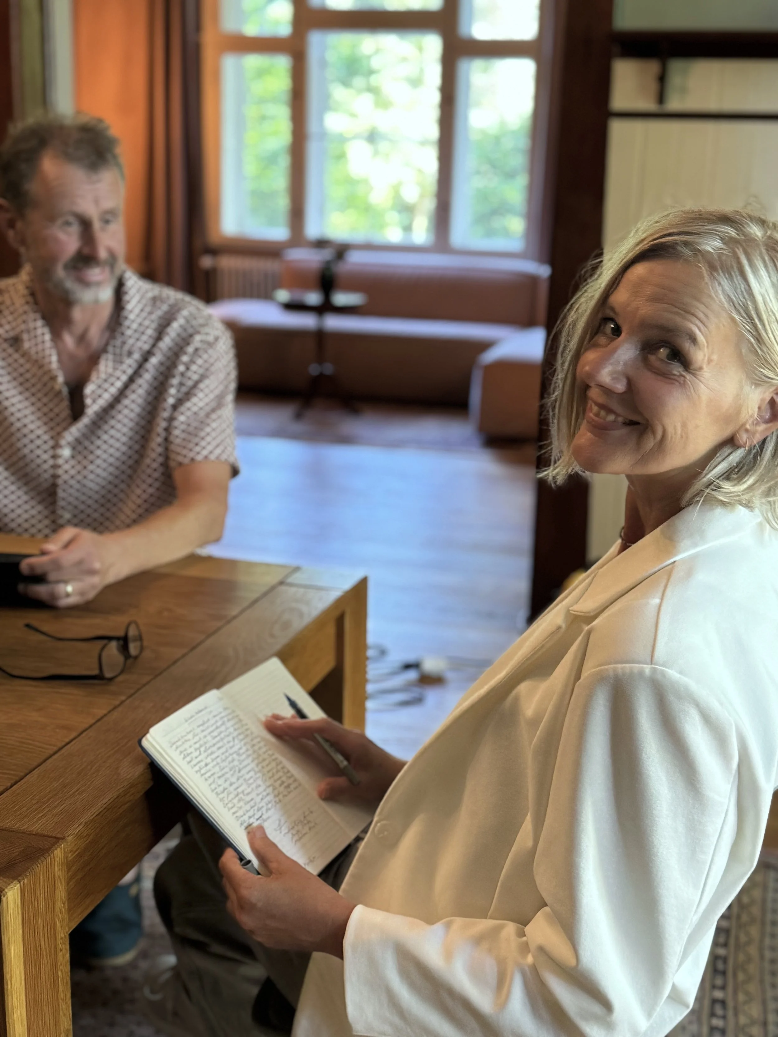 Eine Frau mit einem Notizbuch in der Hand schaut in die Kamera, während ein Mann am Tisch sitzt und zuschaut, in einem Raum mit großen Fenstern und Holzfußboden.