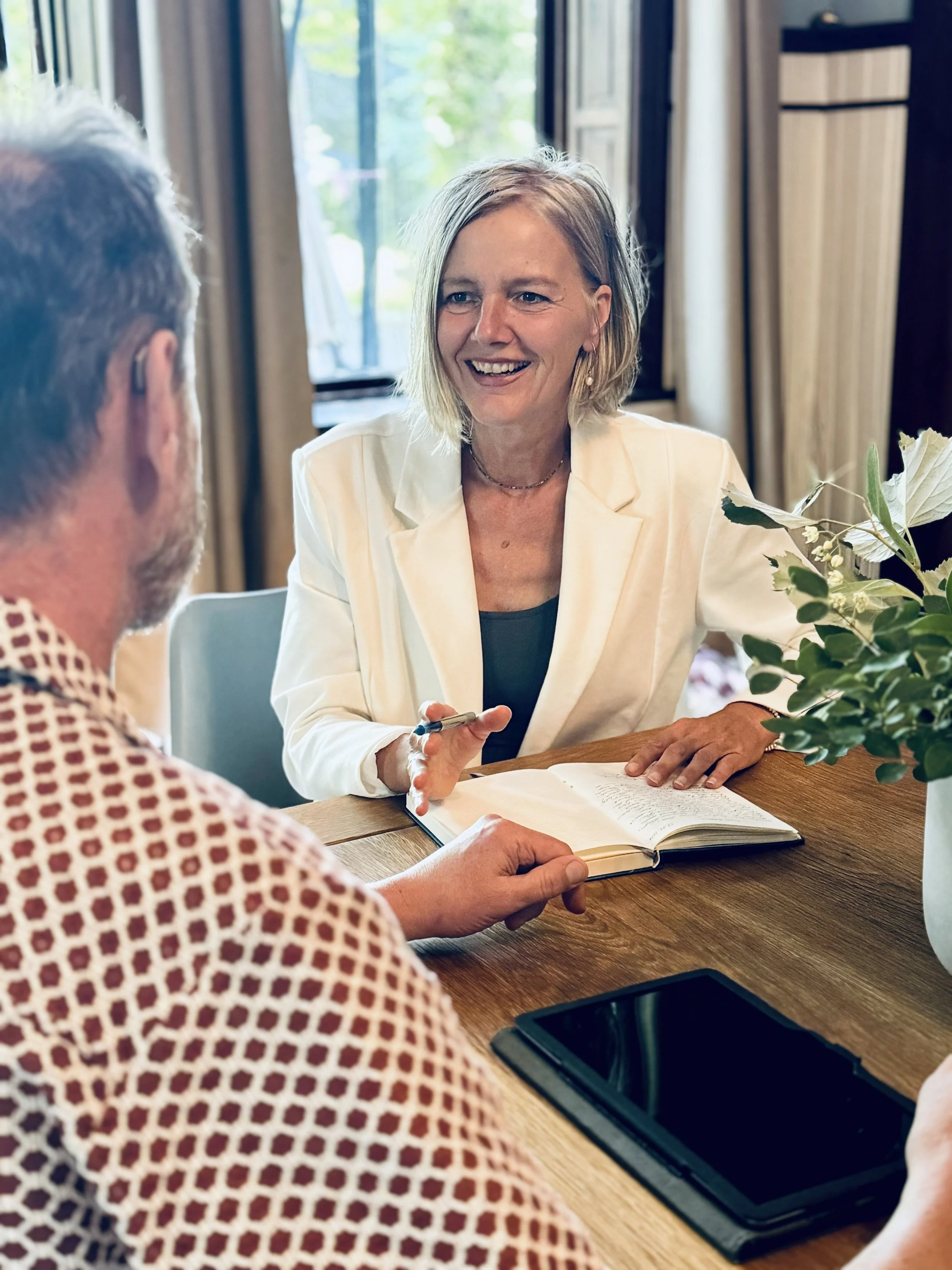 Zwei Menschen sitzen an einem Tisch, eine Frau im weißen Blazer und ein Mann mit grauem Haar, im Hintergrund ein Fenster mit Blick auf Bäume, Tasse und Smartphone auf dem Tisch.