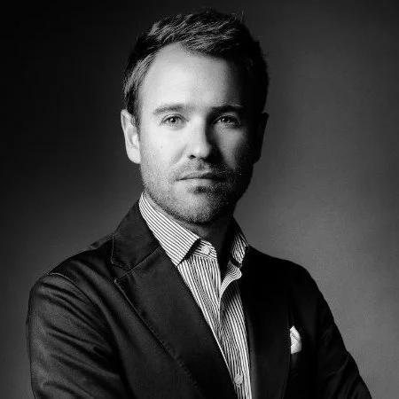 Black and white portrait of a confident man with short dark hair, wearing a blazer over a striped dress shirt.