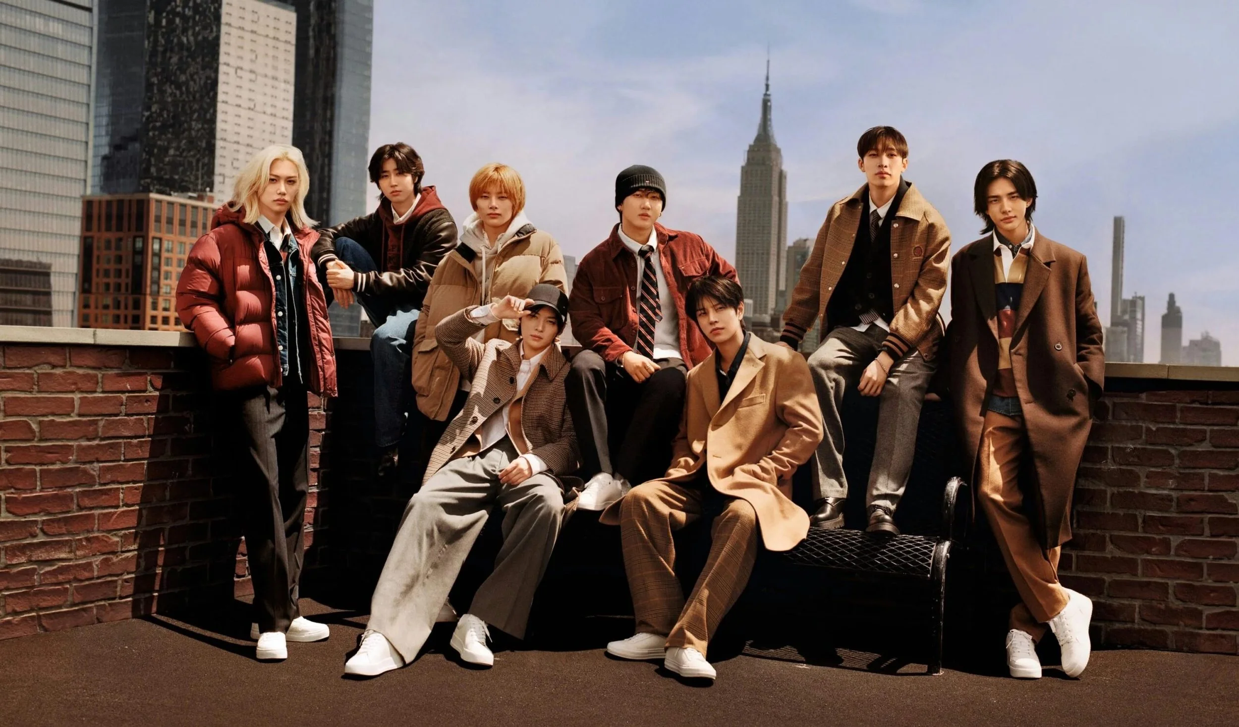 A group of nine young people posing outdoors on a city rooftop, with tall skyscrapers, including the Empire State Building, in the background on a clear day.