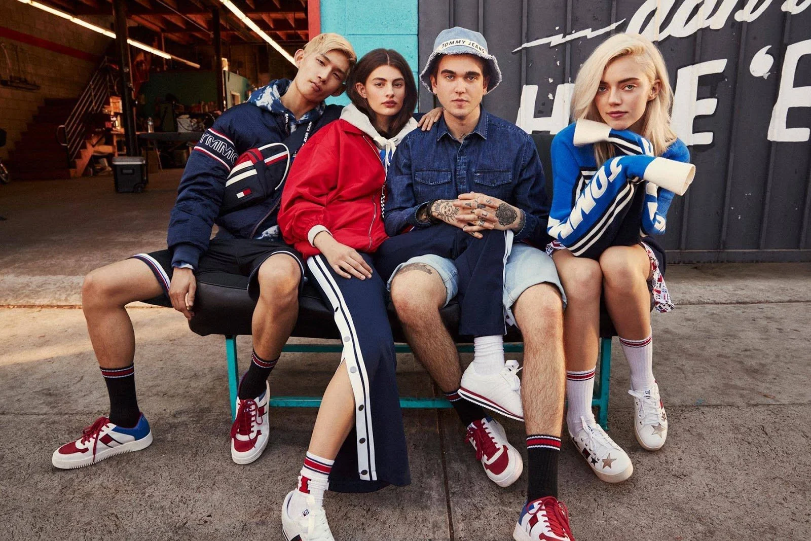 Four young people sitting together on a bench outside an urban building, wearing casual sporty clothing and sneakers, with a graffiti-covered wall behind them.