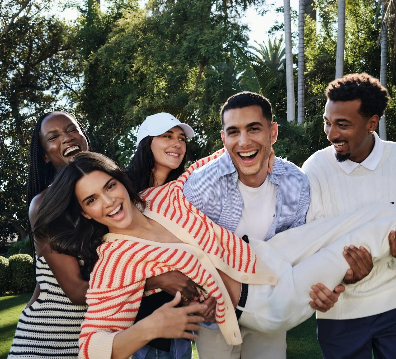A group of five friends outdoors, smiling and playing together on a sunny day with trees in the background.
