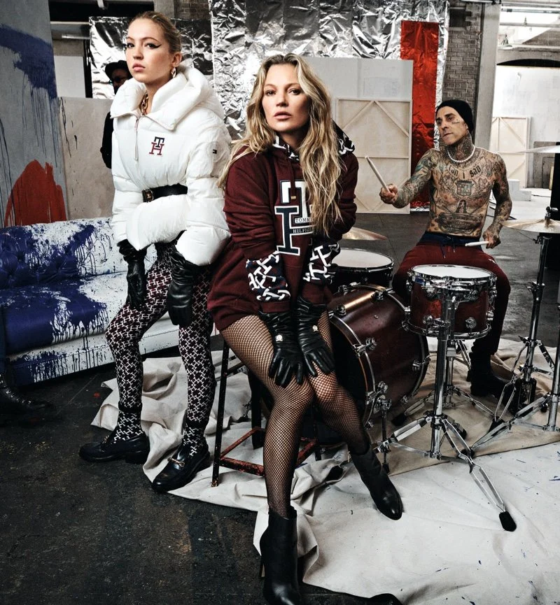 Three models in a photoshoot studio with graffiti, fabric, and industrial backdrops. Two women in fashionable outfits, one seated in front of a drum set, and one standing with her arms crossed, the third person playing drums in the background.