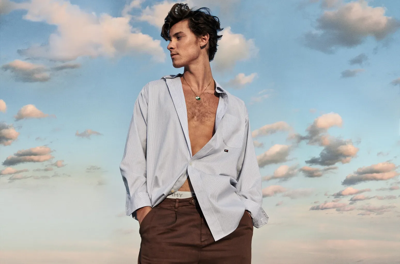 A person standing outdoors under a partly cloudy sky, wearing an unbuttoned, light-colored, striped shirt with a small logo on the chest, brown pants, with hands in pockets, and a necklace.