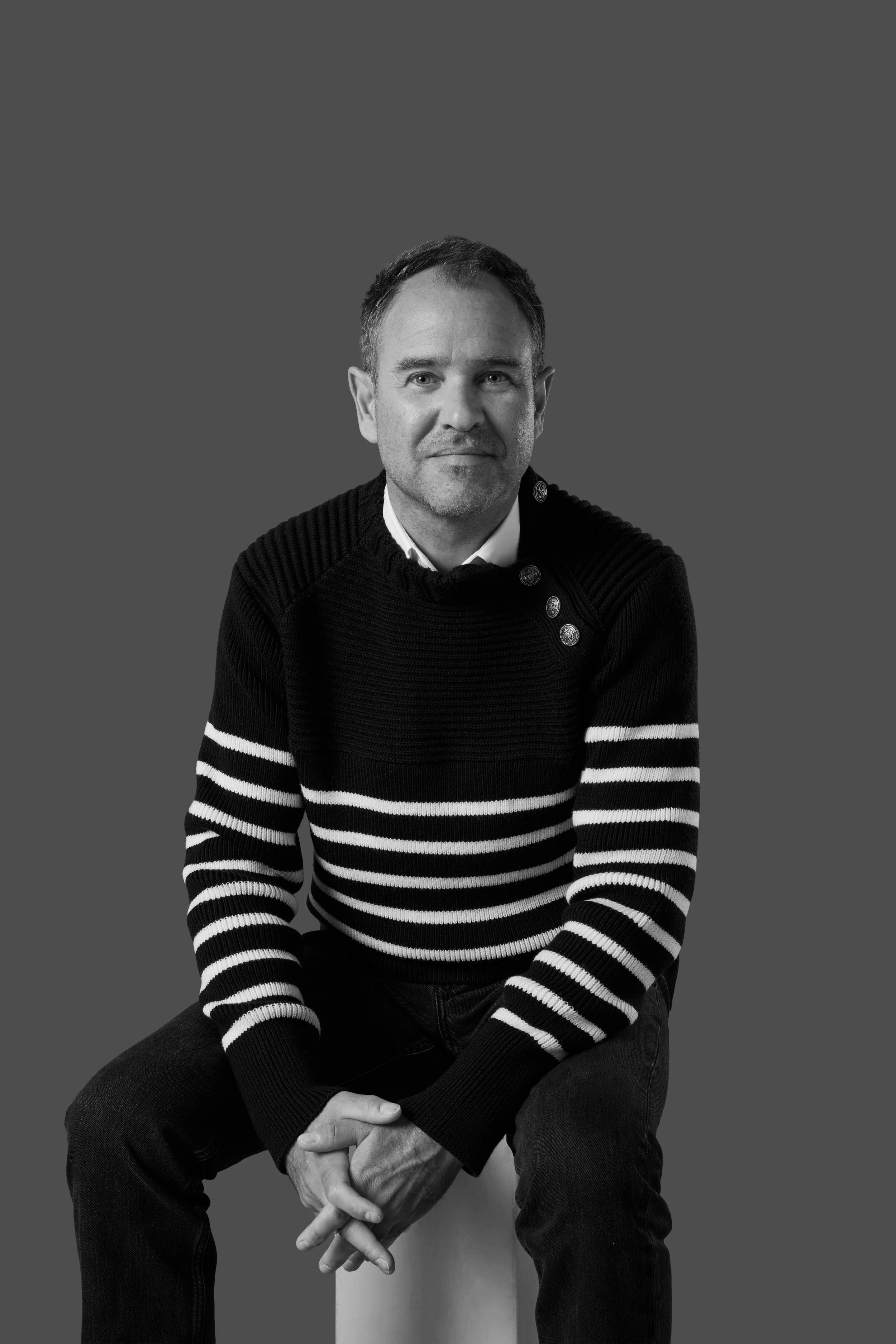 A black and white portrait of a middle-aged man with short hair, sitting against a plain background, wearing a black and white striped sweater with gold buttons on the shoulder, and looking directly at the camera with a optimistic expression.