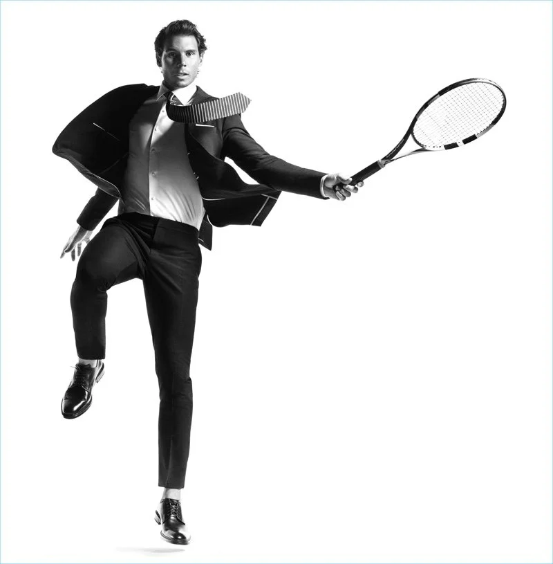 Black and white photo of a man in a suit holding a tennis racket, mid-action pose as if playing tennis.