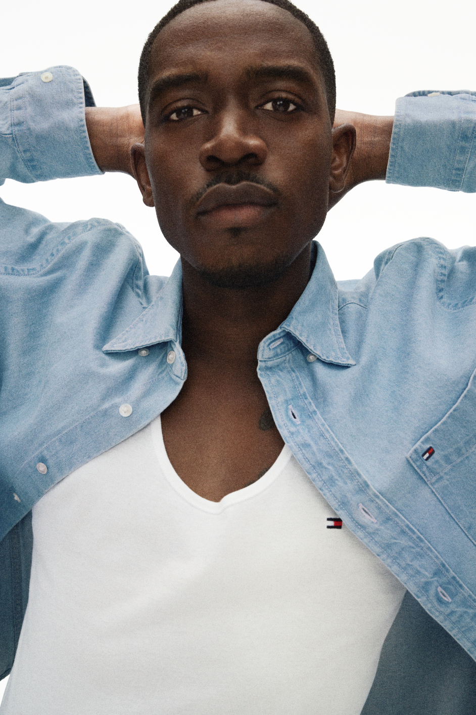 A young Black man with short hair, wearing a light blue denim shirt over a white T-shirt, standing against a plain white background with his arms raised behind his head.