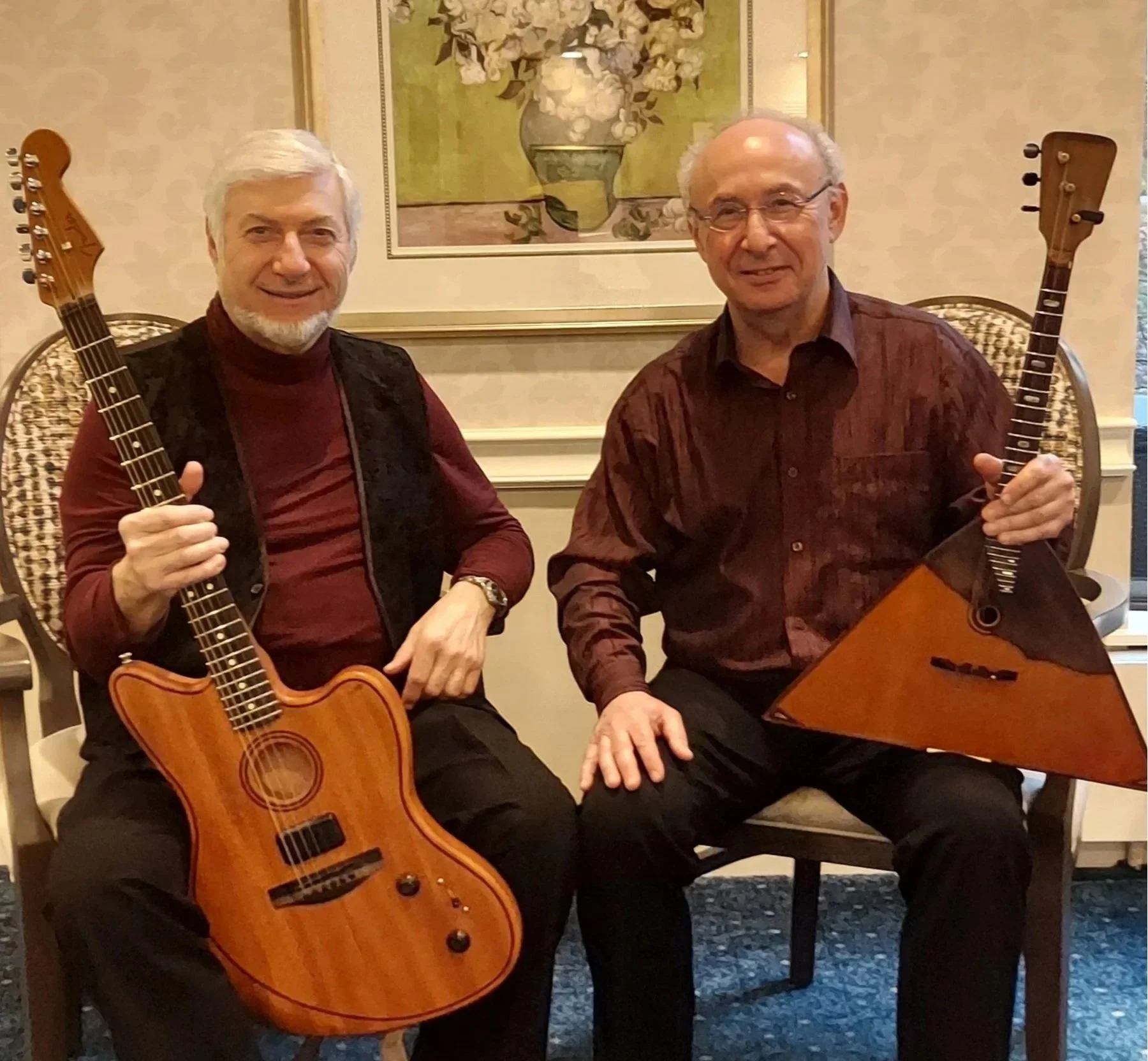 Zwei Männer sitzen im Raum, beide spielen Gitarre. Der Mann auf der linken Seite hat einen grauen Bart und trägt einen dunklen Weste und eine rote Hemd; der Mann auf der rechten Seite ist älter, trägt Brille und ein rotes Hemd. Im Hintergrund ist ein