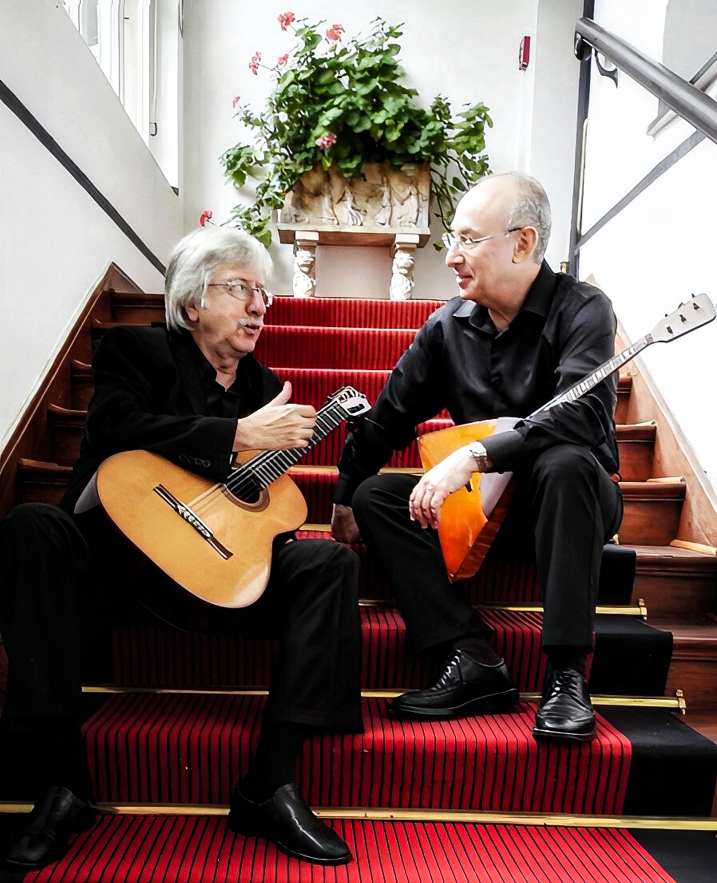 Zwei Männer sitzen in einem Treppenhaus. Der ältere Mann spielt Gitarre und der jüngere Mann hält ein Akkordeon. Sie sprechen miteinander, während sie sich auf den Stufen befinden, im Hintergrund eine große Topfpflanze auf einem Brunst. Ein roter Tep