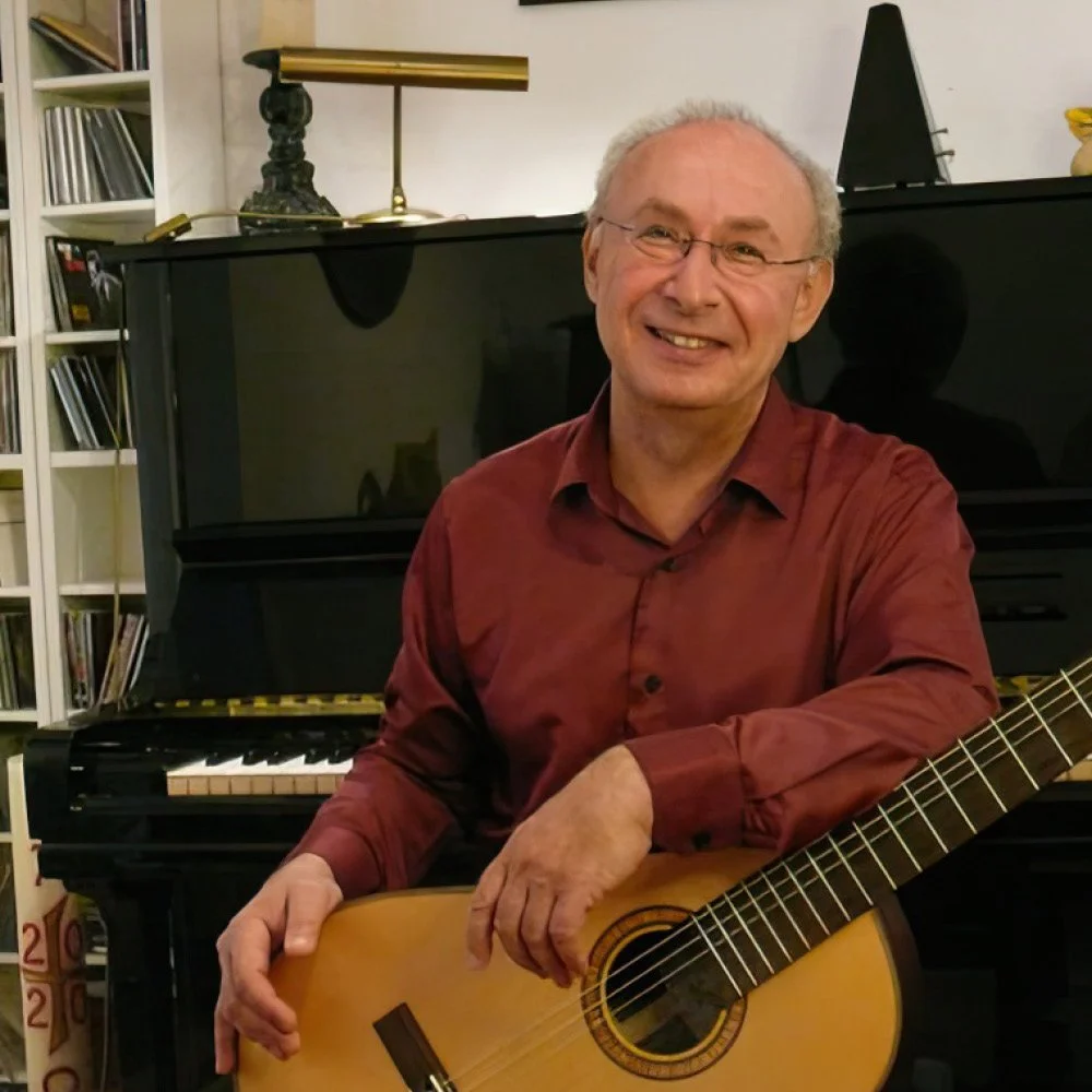Ein lächelnder älterer Mann mit Brille sitzt neben einer Gitarre vor einem schwarzen Klavier in einem Wohnzimmer.