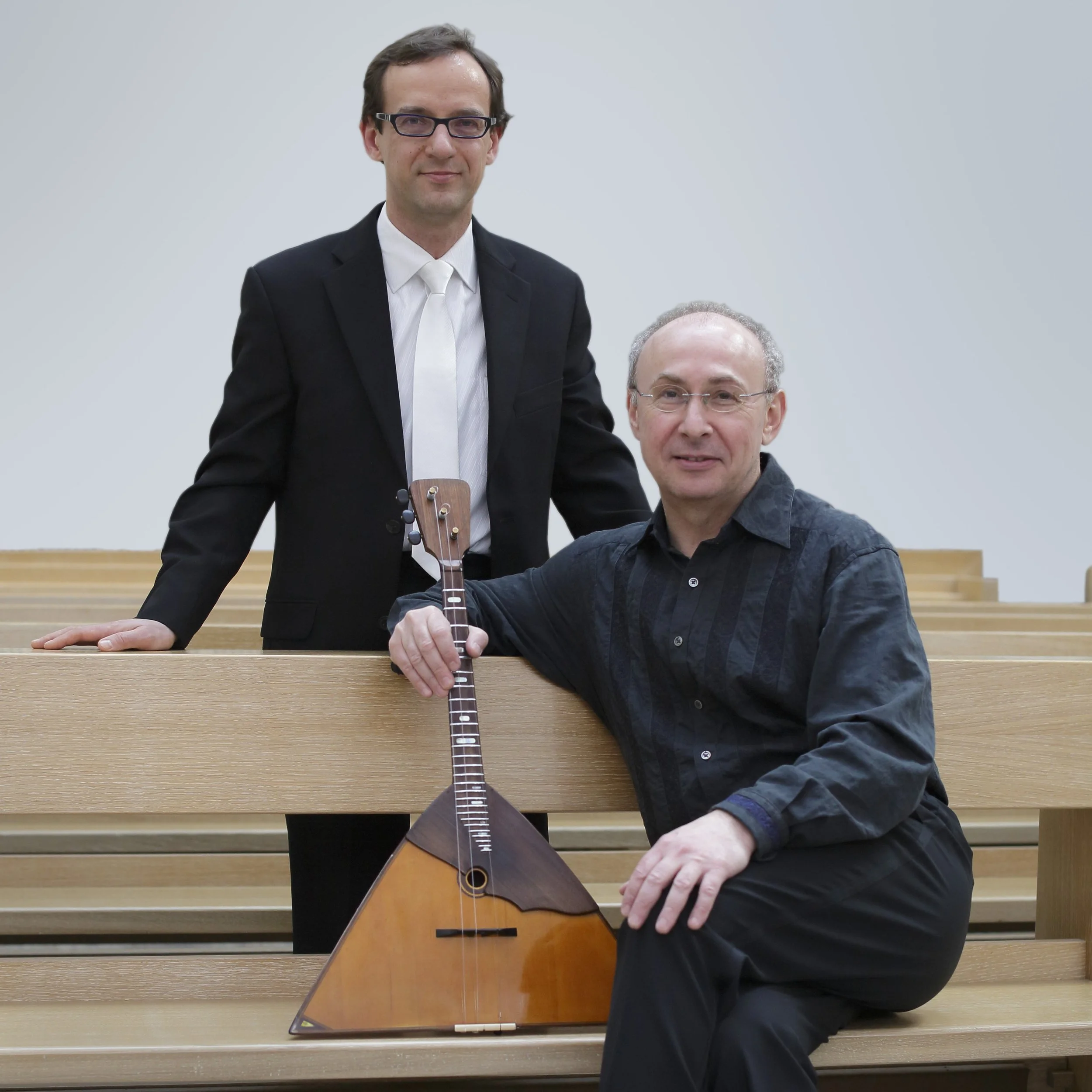 Zwei Männer, einer mit dunklen Haaren und Brille, der andere mit kurzen grauen Haaren, sitzen an einem Holzsamtk, der eine Gitarre hält, vor einem weißen Hintergrund.