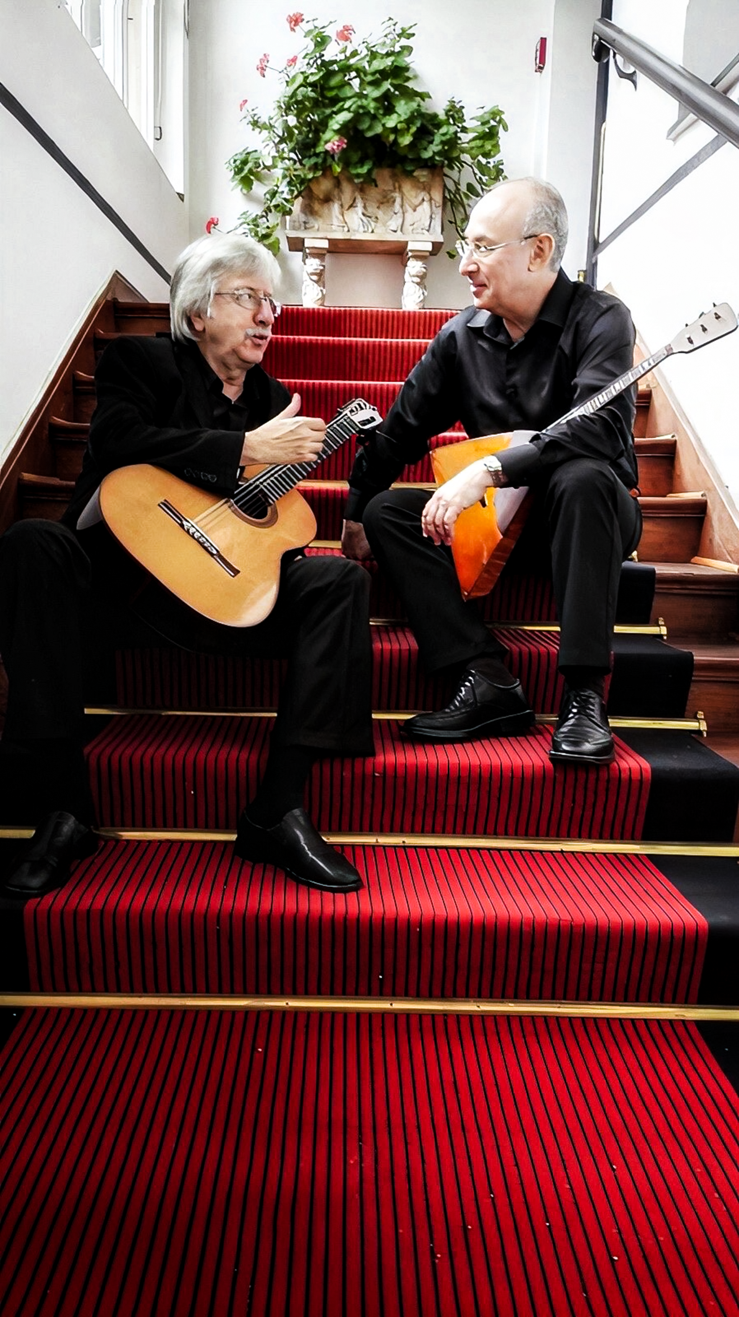 Zwei Männer sitzen auf einer roten Treppe, einer spielt Gitarre, beide unterhalten sich. Im Hintergrund eine Topfpflanze, daneben eine treppenähnliche Einrichtung und ein Fenster.