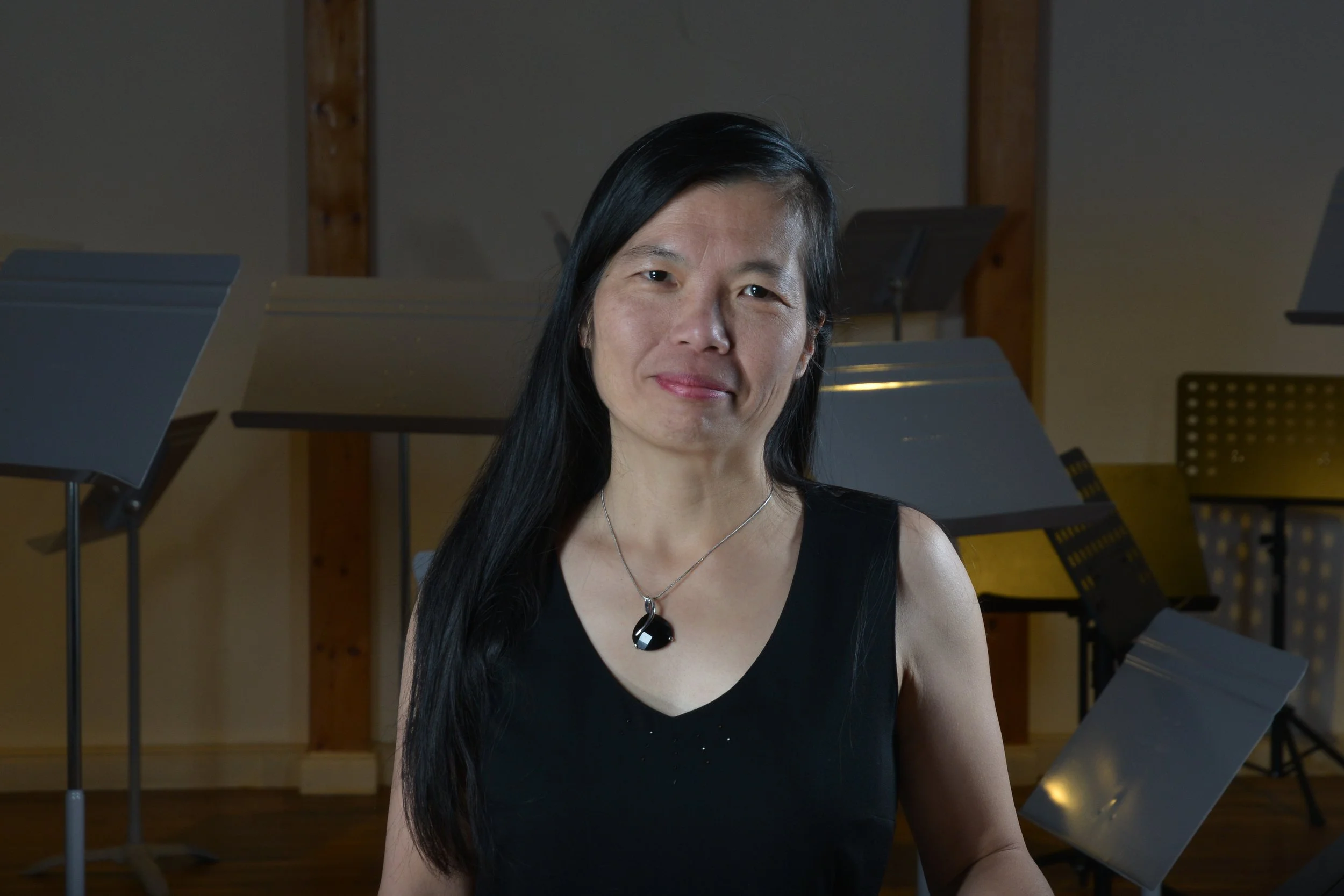Porträt einer asiatisch aussehenden Frau mit langen schwarzen Haaren, schwarzen Kleid und einem schwarzen Schmuck vor einem Raum mit Notenständern und Stühlen.