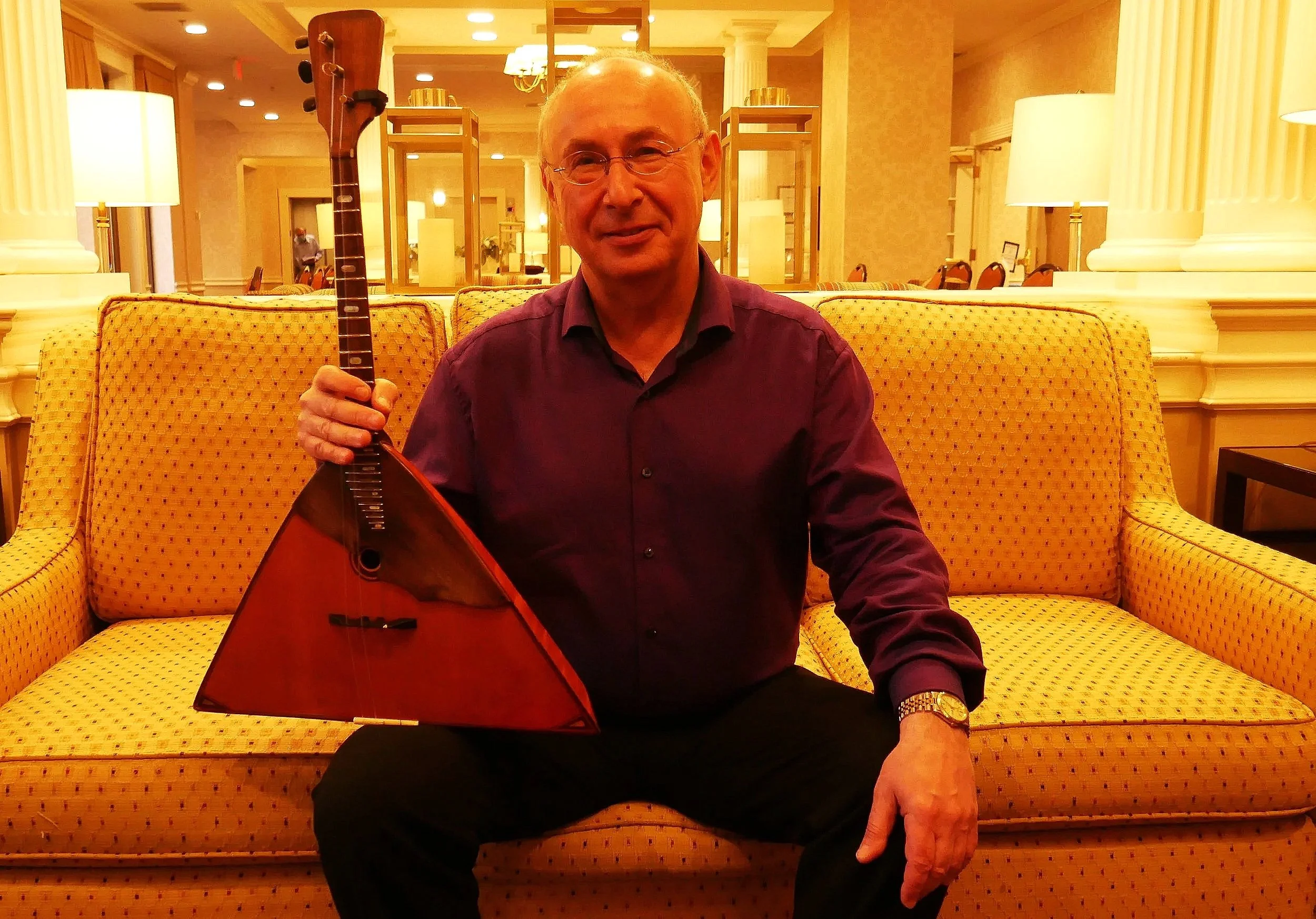 Ein älterer Mann mit Brille und einem Schwarzen Hemd sitzt auf einem gelb-karierten Sofa in einem Hotel-Lobby, hält eine Gitarre und lächelt.