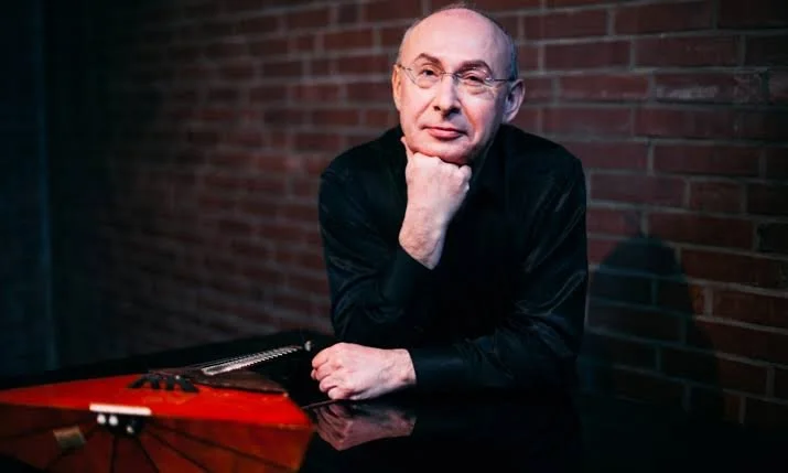 Ein älterer Mann mit Glatze und Brille sitzt an einem Tisch mit einem rot-schwarzen Musikinstrument vor einem Ziegelsteinwand.