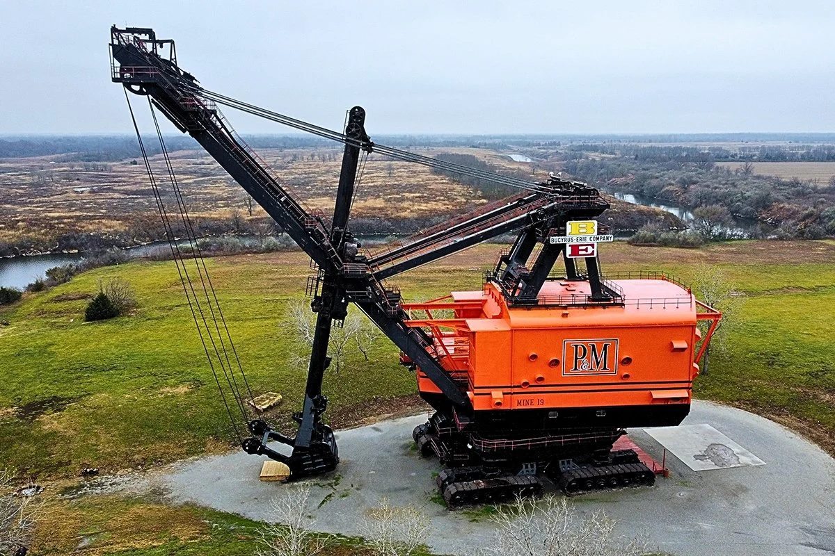A large, orange industrial excavator with a long arm and cables, situated on a gravel surface in an open field.