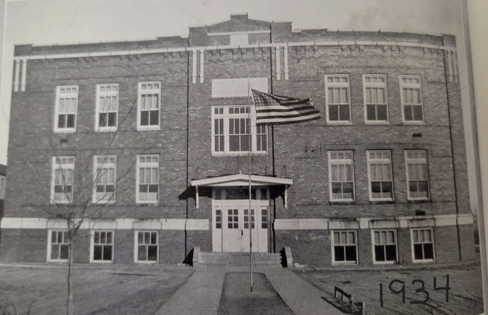 Black and white photo of a brick school building with multiple windows, a central entrance, steps leading up to the door, and an American flag hanging beside the entrance. The year 1934 is written on the bottom right corner.