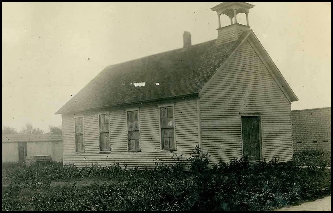 An old black-and-white photo of a small wooden church with a pitched roof, four windows, a door on the front, and a small bell tower on top, surrounded by low bushes.