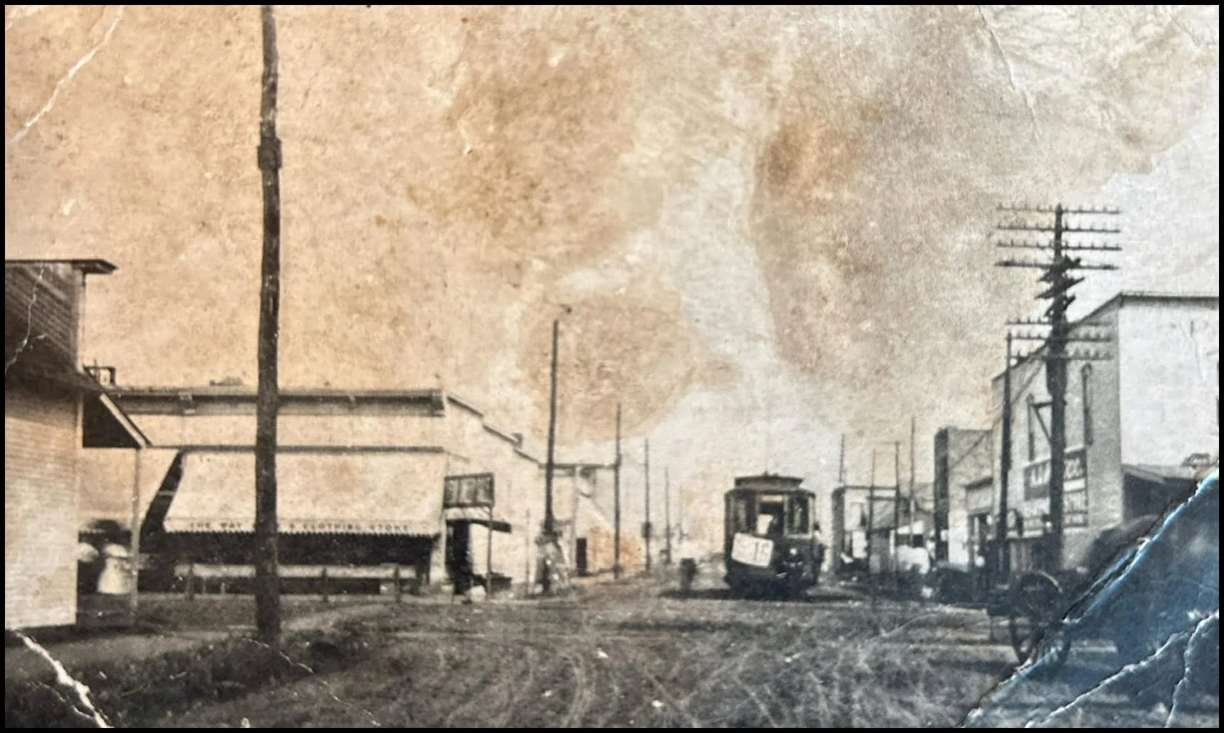 An old black-and-white photograph of a street scene with buildings, utility poles, and a streetcar or tram on a dirt road.