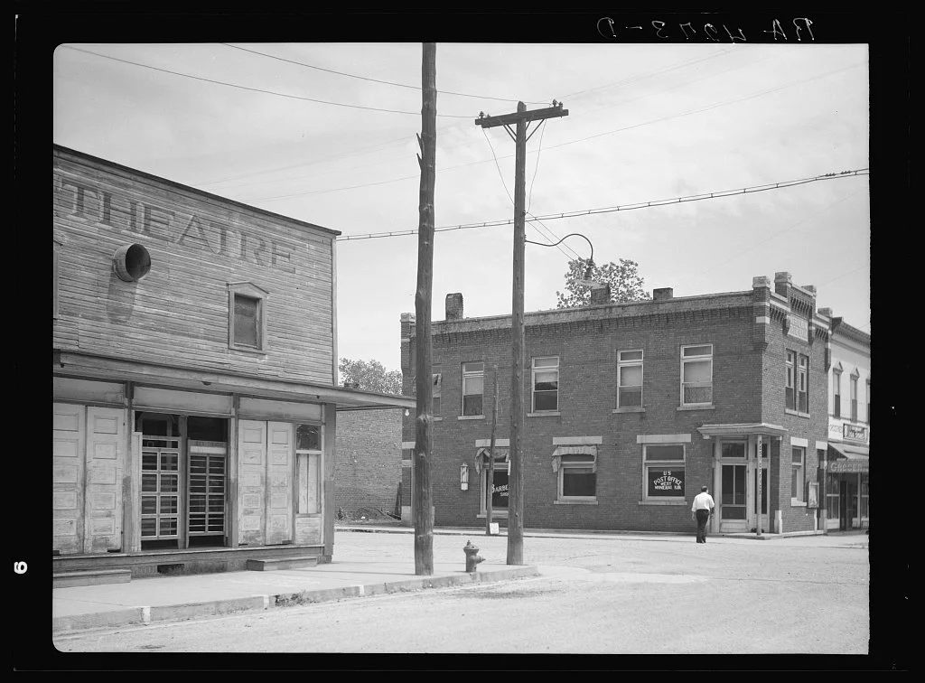 Black and white photo of a small town intersection with old brick buildings, utility poles, and a person walking on the sidewalk.