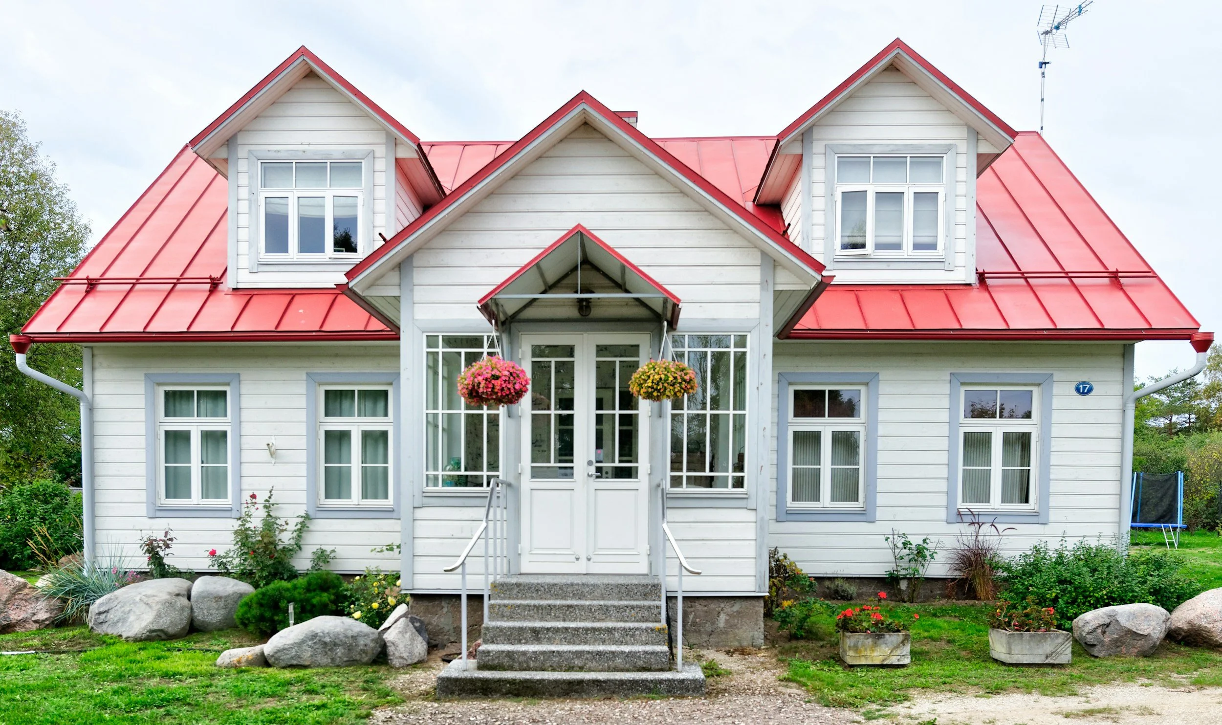 A two-story white house with a red metal roof, multiple large windows, a small porch with stairs, hanging flower baskets, and a garden with rocks and plants in front.
