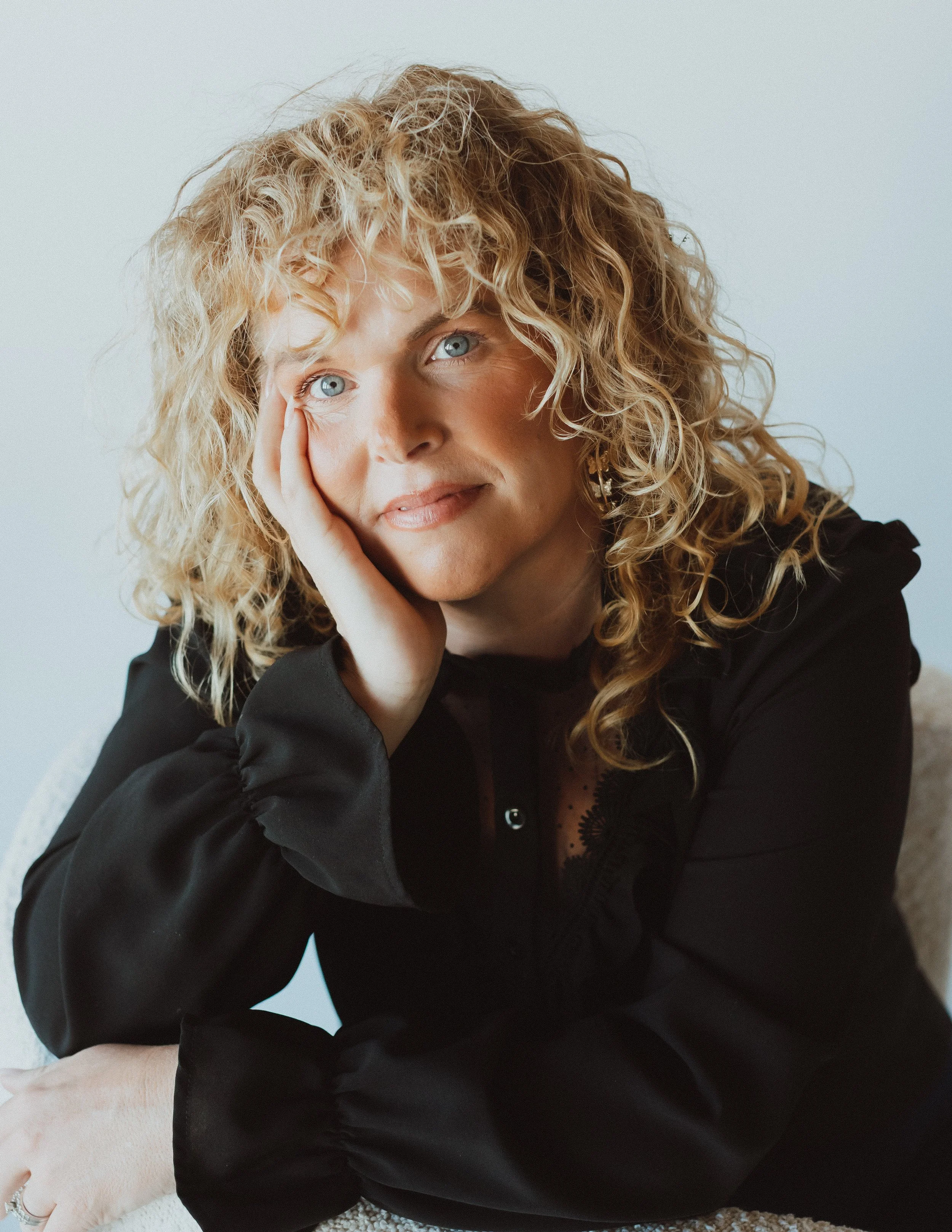 A woman with curly blonde hair resting her head on her hand, wearing a black top, and sitting against a neutral background.