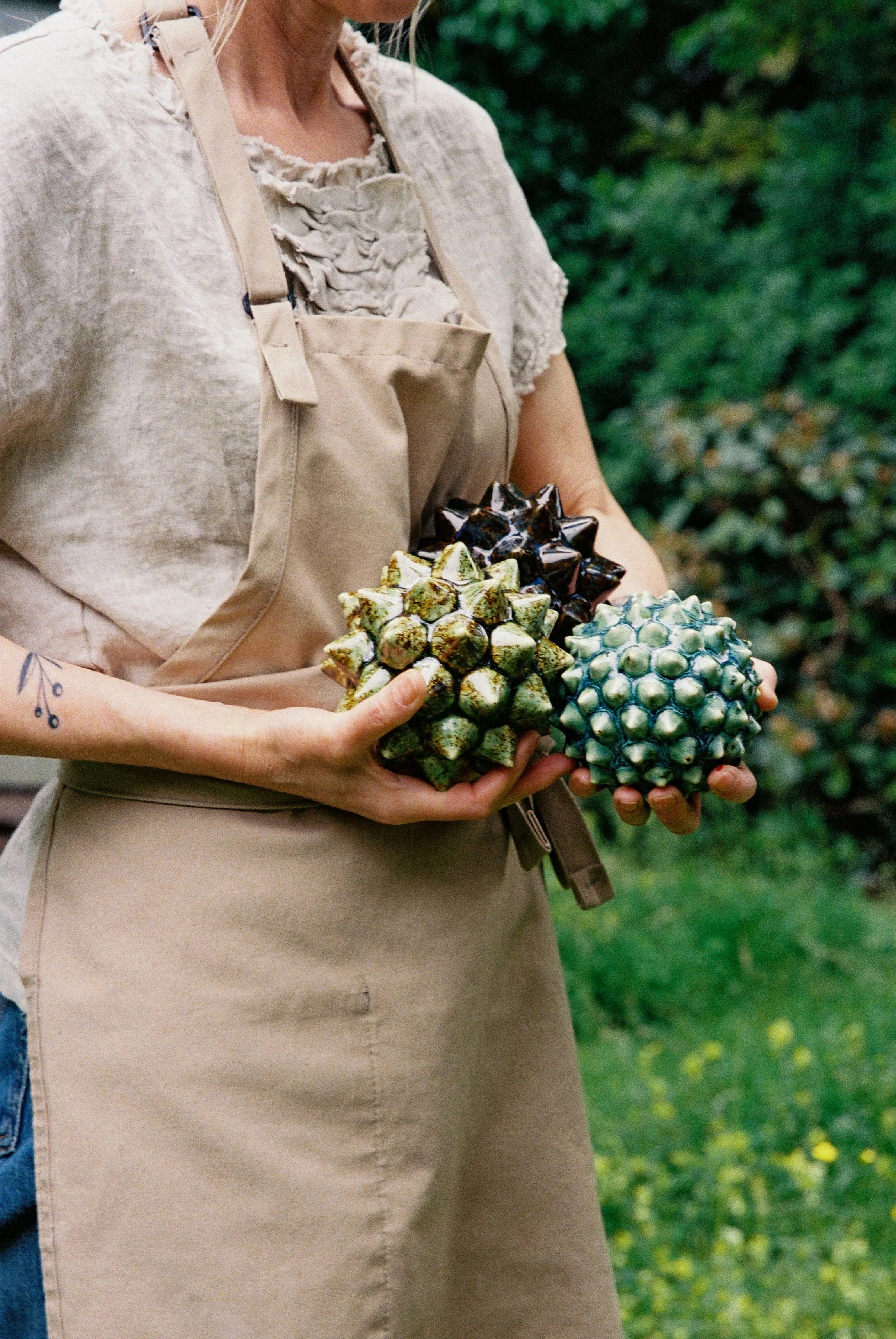 A person wearing a beige apron holding three ceramic art pieces shaped like spiky, textured balls, with a background of green foliage.